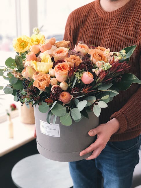 A person in a rust-colored knitted sweater is holding a large, round grey floral hat box with a white label reading 'Secret Garden.' The box contains a vibrant bouquet of fresh flowers, including peac
