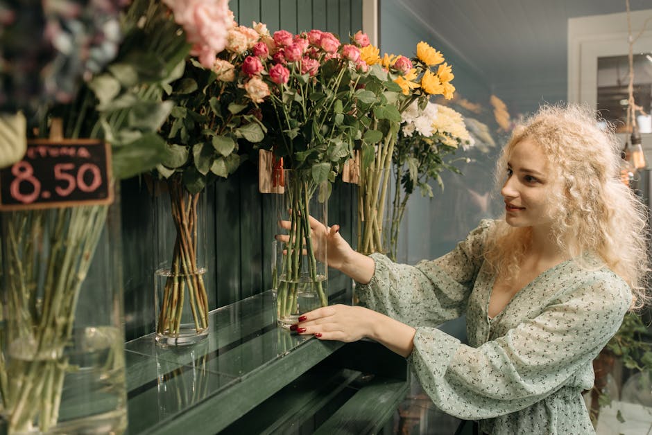 A young woman with curly blonde hair and fair skin arranging a bouquet of fresh flowers at a florist shop. She is wearing a light green, flowery blouse and has red painted nails. The bouquet features 
