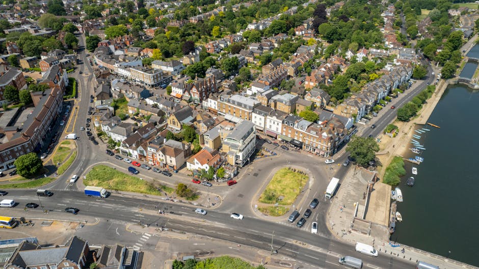 Aerial view of Bounds Green featuring a cluster of residential buildings, shops, and a waterside area with boats docked along the shoreline. The scene includes a main road with moving vehicles, small
